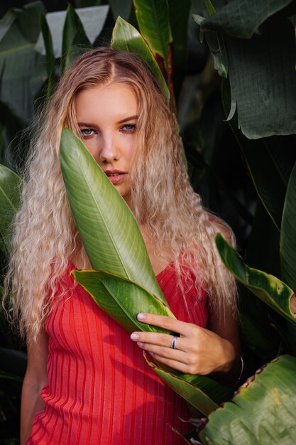 Kostenloses Foto porträt der jungen kaukasischen frau im roten passenden eleganten kleid im freien auf hintergrund der tropischen blätter