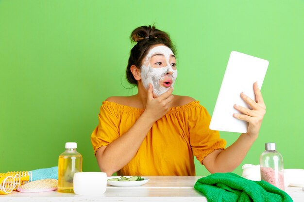 Porträt der jungen kaukasischen Frau im Schönheitstag, Haut- und Haarpflege-Routine. Weibliches Modell, das Selfie, Vlog oder Videoanruf macht, während Gesichtsmaske angewendet wird. Selbstpflege-, Naturschönheits- und Kosmetikkonzept.