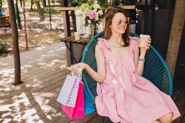 Porträt der jungen lächelnden glücklichen attraktiven Frau, die im Café mit Einkaufstüten sitzt, die Kaffee, Sommermode-Outfit, rosa Baumwollkleid, trendige Kleidung trinken