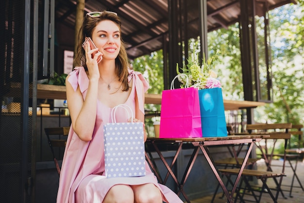 Porträt der jungen lächelnden glücklichen attraktiven Frau, die im Café sitzt und am Telefon mit Einkaufstaschen, Sommermode-Outfit, Hipster-Stil, rosa Baumwollkleid, trendige Kleidung spricht