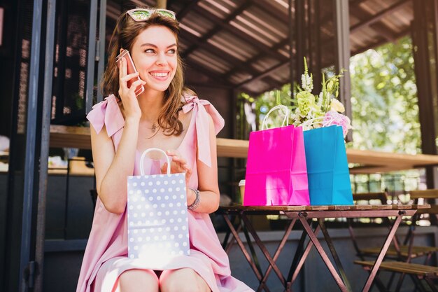 Porträt der jungen lächelnden glücklichen attraktiven Frau, die im Café sitzt und am Telefon mit Einkaufstaschen, Sommermode-Outfit, Hipster-Stil, rosa Baumwollkleid, trendige Kleidung spricht