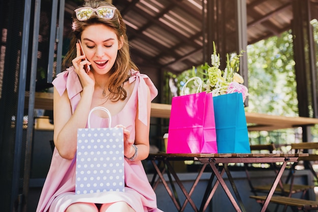 Porträt der jungen lächelnden glücklichen attraktiven Frau, die im Café sitzt und am Telefon mit Einkaufstaschen, Sommermode-Outfit, rosa Baumwollkleid, überraschtes Gesicht spricht