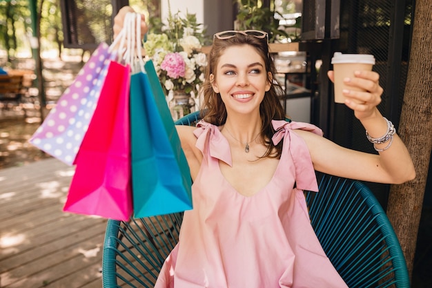 Porträt der jungen lächelnden glücklichen hübschen Frau mit aufgeregtem Gesichtsausdruck, der im Café mit Einkaufstüten sitzt, die Kaffee, Sommermode-Outfit, rosa Baumwollkleid, trendige Kleidung trinken