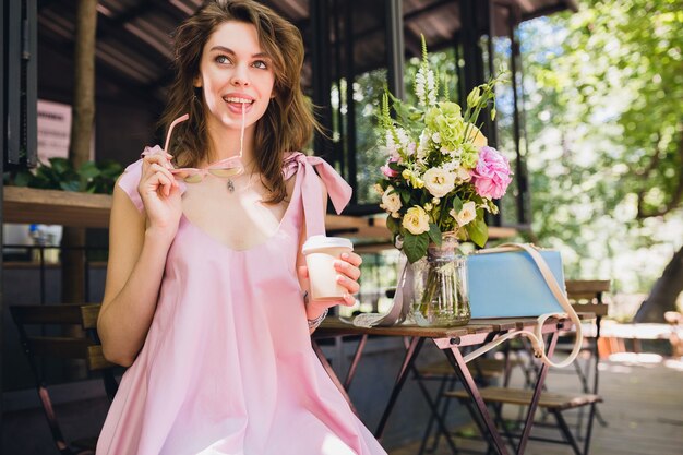 Porträt der jungen lächelnden glücklichen hübschen Frau mit dem Sitzen im Café, das Kaffee trinkt, Sommermode-Outfit, Hipster-Stil, rosa Baumwollkleid, trendige Bekleidungszubehör