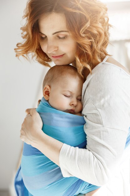 Porträt der jungen Mutter und des neugeborenen Sohnes, die auf Mutterbrust in der blauen Babyschlinge schlafen. Familienglücksstimmung. Familienkonzept.