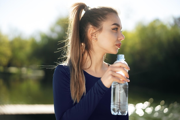 Porträt der jungen schönen Frau, die blaues Sportkleidungstrinkwasser im Park trägt