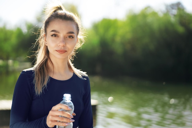 Porträt der jungen schönen Frau, die blaues Sportkleidungstrinkwasser im Park trägt