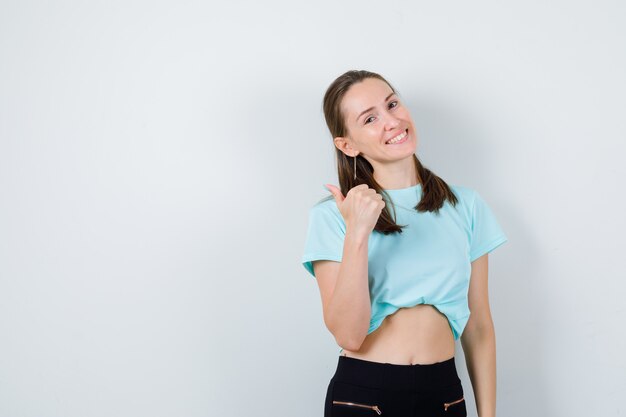 Porträt der jungen schönen Frau, die mit dem Daumen im T-Shirt zurück zeigt und fröhliche Vorderansicht schaut