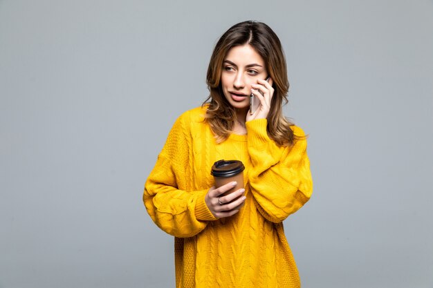 Porträt der jungen schönen Frau in der gelben Spitze, die Pappbecher des Kaffee zum Mitnehmen hält und glücklich lokalisiert auf grauer Wand lächelt