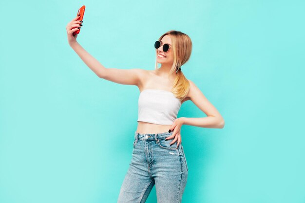 Porträt der jungen schönen lächelnden blonden Frau in trendiger Sommerkleidung Sexy sorglose Frau posiert in der Nähe der blauen Wand im Studio Positives Modell, das sich drinnen amüsiert Fröhlich und glücklich Selfie nehmen