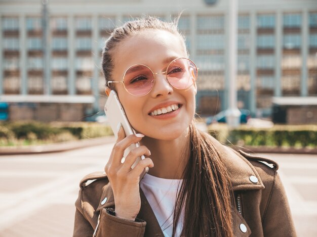 Porträt der jungen schönen lächelnden Frau, die am Telefon spricht Modisches Mädchen in der zufälligen Sommerkleidung Lustige und positive Frau, die auf der Straße aufwirft