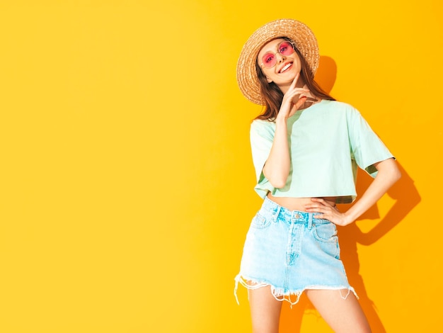 Porträt der jungen schönen lächelnden Frau im trendigen Sommerjeansrock sorglose Frau, die in der Nähe der gelben Wand im Studio posiert Positives Modell, das sich drinnen amüsiert Fröhlich und glücklich In Hut