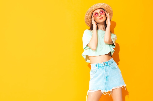 Porträt der jungen schönen lächelnden Frau im trendigen Sommerjeansrock sorglose Frau, die in der Nähe der gelben Wand im Studio posiert Positives Modell, das sich drinnen mit Sonnenbrille amüsiert Fröhlich und glücklich In Hut