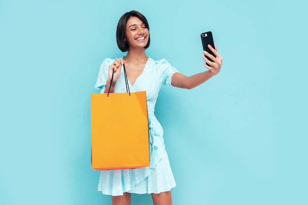 Porträt der jungen schönen lächelnden Frau im trendigen Sommerkleid Sorglose Frau posiert in der Nähe der blauen Wand im Studio Positives Modell mit Einkaufstasche Fröhlich und glücklich Selfie machen
