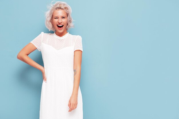 Porträt der jungen schönen lächelnden Frau im trendigen weißen Sommerkleid Sexy sorglose blonde Frau posiert in der Nähe der blauen Wand im Studio Positives Modell, das sich drinnen amüsiert Fröhlich und glücklich