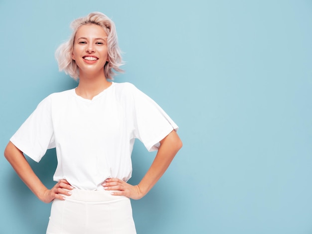 Kostenloses Foto porträt der jungen schönen lächelnden frau im trendigen weißen sommerkleid sexy sorglose blonde frau posiert in der nähe der blauen wand im studio positives modell, das sich drinnen amüsiert fröhlich und glücklich