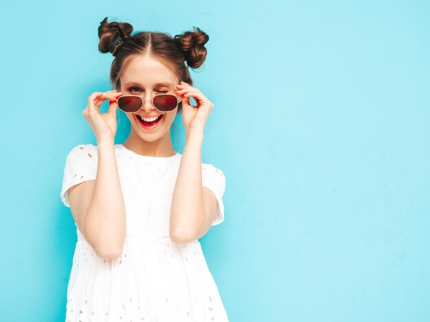 Porträt der jungen schönen lächelnden Frau im trendigen weißen Sommerkleid sorglose Frau posiert in der Nähe der blauen Wand im Studio Positives Modell, das sich drinnen amüsiert Fröhlich und glücklich Nimmt Sonnenbrille ab