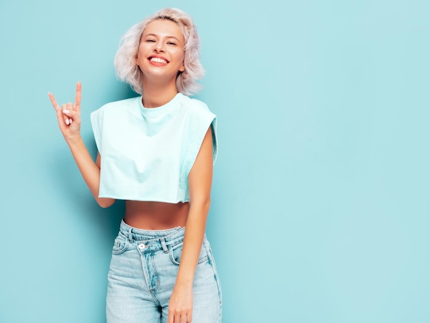 Porträt der jungen schönen lächelnden Frau in trendiger Sommerkleidung Sexy sorglose Frau posiert in der Nähe der blauen Wand im Studio Positives blondes Modell, das Spaß hat und verrückt wird Zeigt Rock-and-Roll-Zeichen