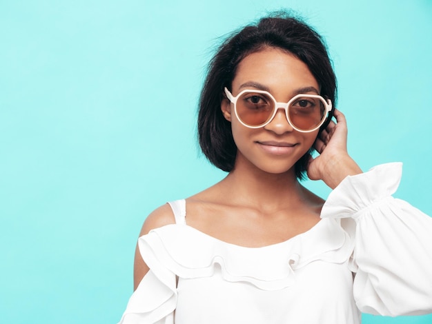 Porträt der jungen schönen schwarzen Frau Lächelndes Modell in Sommerjeans gekleidet Sexy sorglose Frau posiert in der Nähe der blauen Wand im Studio Gebräunt und fröhlich In Sonnenbrille