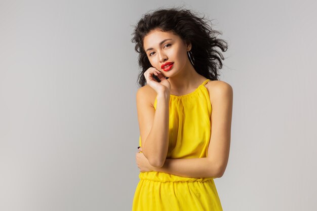 Porträt der jungen sexy brünetten Frau im gelben Kleid, das ihr Gesicht berührend, rote Lippen, lockiges Haar, das im Wind weht, Sommerart, Modetrend, Blick in die Kamera, gemischte Rasse, isoliert, Händchen haltend