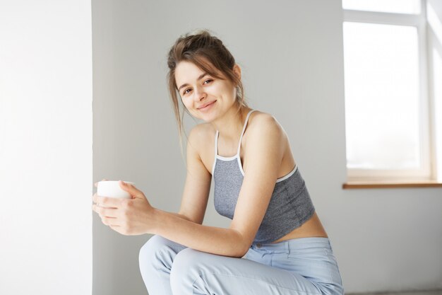 Porträt der jungen zarten Frau lächelnd halten Tasse, die auf Stuhl über weißer Wand am frühen Morgen sitzt.