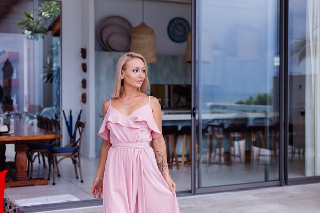 Porträt der kaukasischen Frau im romantischen eleganten rosa langen Kleid im Urlaub im luxuriösen reichen Villenhotel mit erstaunlicher tropischer Palmenansicht-Frau im klassischen weißen Hut