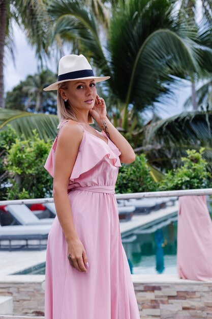 Porträt der kaukasischen Frau im romantischen eleganten rosa langen Kleid im Urlaub im luxuriösen reichen Villenhotel mit erstaunlicher tropischer Palmenansicht-Frau im klassischen weißen Hut