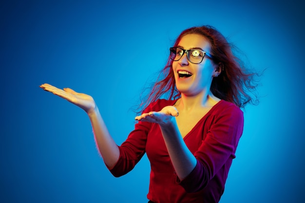 Porträt der kaukasischen Frau lokalisiert auf blauem Studiohintergrund im Neonlicht. Schönes weibliches Modell mit roten Haaren in lässig. Konzept der menschlichen Emotionen, Gesichtsausdruck, Verkauf, Anzeige. Gruß, geschockt.