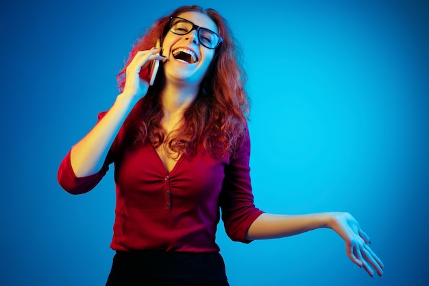 Porträt der kaukasischen Frau lokalisiert auf blauem Studiohintergrund im Neonlicht. Schönes weibliches Modell mit roten Haaren in lässig. Konzept der menschlichen Emotionen, Gesichtsausdruck, Verkauf, Anzeige. Telefonieren.