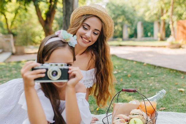 Porträt der lachenden langhaarigen Dame im Hut mit Mädchen, das Kamera hält. Außenfoto der jungen Frau, die Spaß am Picknick und ihrer Tochter hat, die besie Korb mit Mahlzeit sitzt.