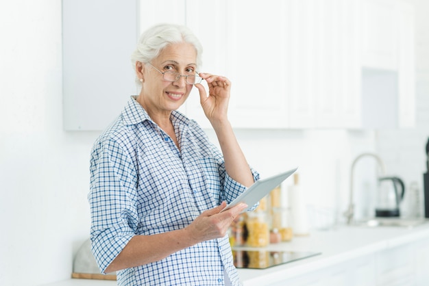 Porträt der lächelnden älteren Frau, welche die digitale Tablette steht in der Küche hält