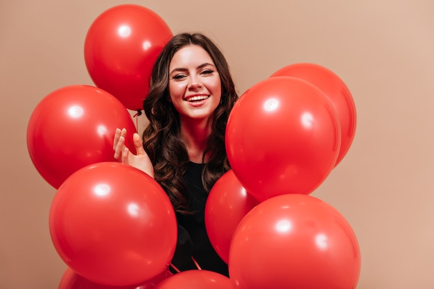 Porträt der lächelnden Frau mit welligem dunklem Haar, das mit riesigen Luftballons auf beigem Hintergrund aufwirft.