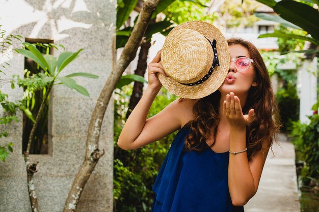 Porträt der lächelnden glücklichen attraktiven jungen Frau im blauen Kleid und im Strohhut, die rosa Sonnenbrillen tragen, die im tropischen Spa-Villenhotel im Urlaub im Sommerart-Outfit gehen