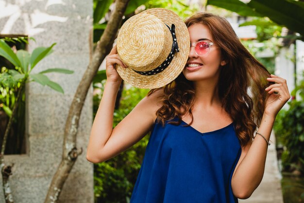 Porträt der lächelnden glücklichen attraktiven jungen Frau im blauen Kleid und im Strohhut, die rosa Sonnenbrillen tragen