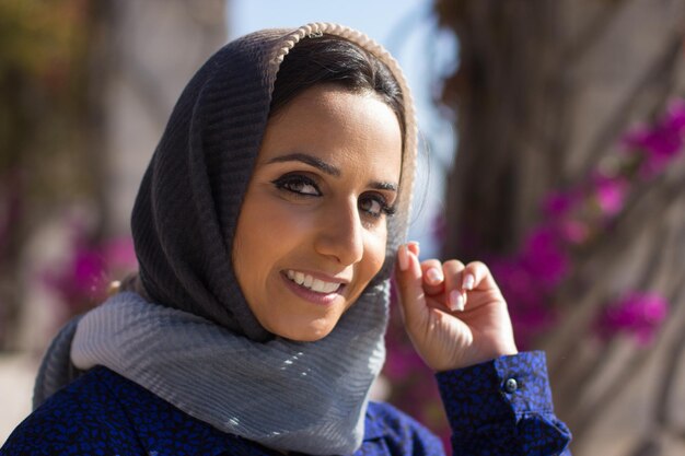 Porträt der lächelnden jungen arabischen Frau. Schöne Frau, die draußen in der Sonne steht und leicht ihr graues Kopftuch berührt. Schönheits- und Ethnizitätskonzept