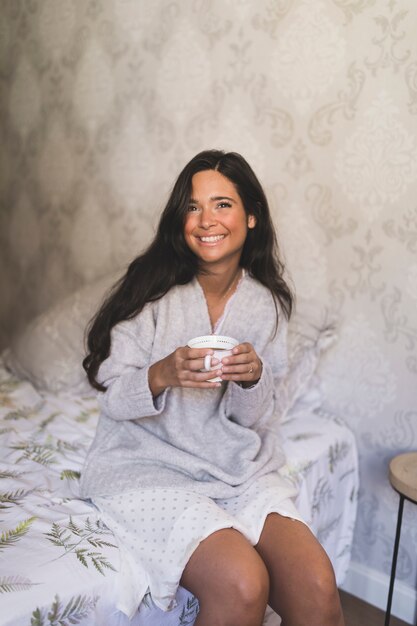 Porträt der lächelnden jungen Frau, die auf dem Bett hält Kaffeetasse sitzt