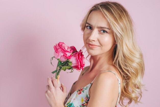 Porträt der lächelnden jungen Frau, die rosa Rosen gegen rosa Hintergrund hält