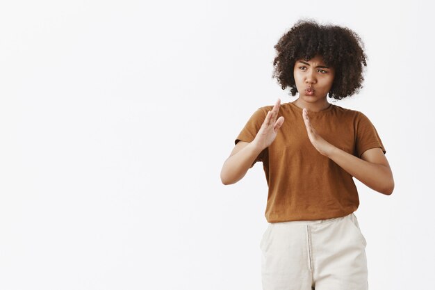 Porträt der lustigen und niedlichen verspielten afroamerikanischen Frau mit den lockigen Haaren im trendigen T-Shirt, das Palmen in Kung-Fu-Pose hebt, faltet Lippen über graue Wand