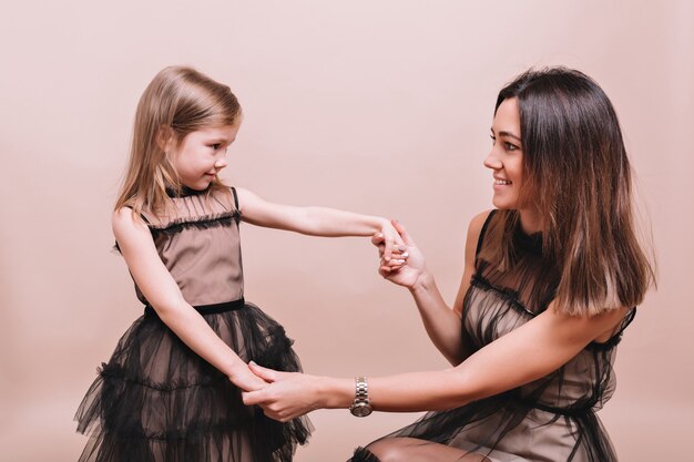 Porträt der modischen jungen Frau mit dem kleinen niedlichen Mädchen, das ähnliche schwarze Kleider trägt, die auf beige Wand mit wahrhaft Emotionen aufwerfen. Stilvoller Familienlook von Mutter und Tochter