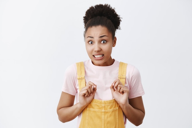 Porträt der nervösen niedlichen afroamerikanischen Frau in gelben Overalls, bückenden und zusammengebissenen Zähnen, entschuldigend und wegnehmend, besorgt über graue Wand stehend