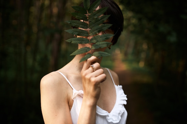 Porträt der nicht wiedererkennbaren mysteriösen jungen frau, die weißes riemenkleid trägt, das im wald allein das gesicht mit großem farnblatt bedeckend aufwirft