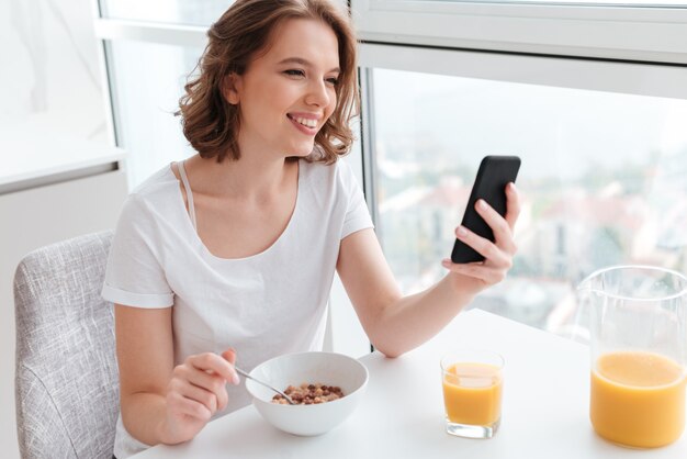 Porträt der niedlichen lächelnden Frau im weißen T-Shirt, das auf Smartphone beim Sitzen und Essen von Cornflakes am Küchentisch plaudert