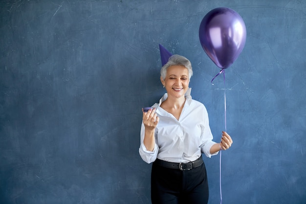 Porträt der positiven sorglosen reifen frau, die geburtstag feiert, glücklichen gesichtsausdruck habend, heliumballon und makrone haltend