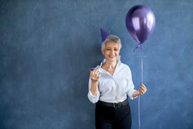 Porträt der positiven sorglosen reifen Frau, die Geburtstag feiert, glücklichen Gesichtsausdruck habend, Heliumballon und Makrone haltend