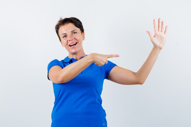 Porträt der reifen Frau, die auf ihre erhobene Hand im blauen T-Shirt zeigt und selbstbewusste Vorderansicht schaut