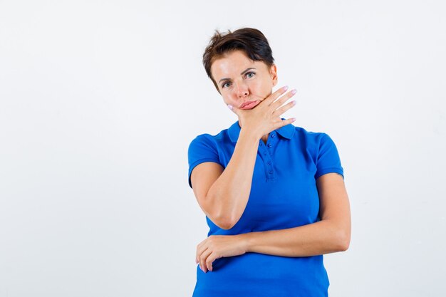 Porträt der reifen Frau, die Hand am Kinn im blauen T-Shirt hält und nachdenkliche Vorderansicht schaut