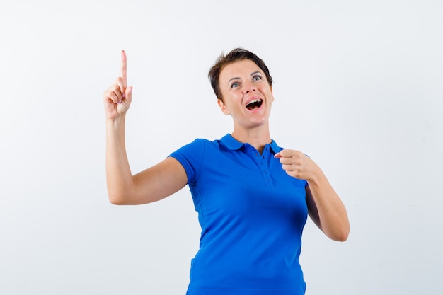 Porträt der reifen Frau, die nach oben zeigt und vorgibt, etwas im blauen T-Shirt zu halten und glückliche Vorderansicht zu schauen