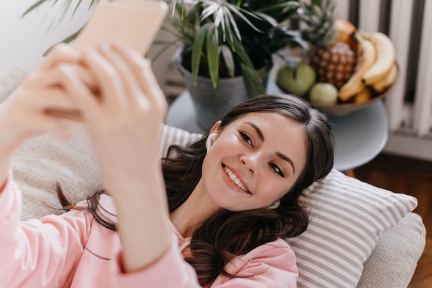 Porträt der reizenden Frau, die auf Kissen im Wohnzimmer liegt und selfie nimmt