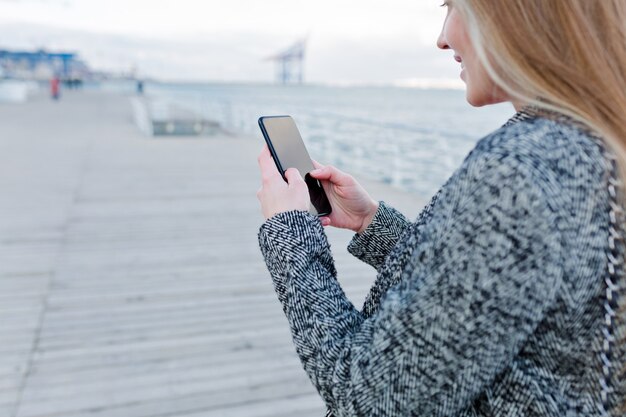 Porträt der reizenden jungen Frau mit glücklichem Lächeln im grauen Mantel, der Smartphone nahe dem Meer rollt