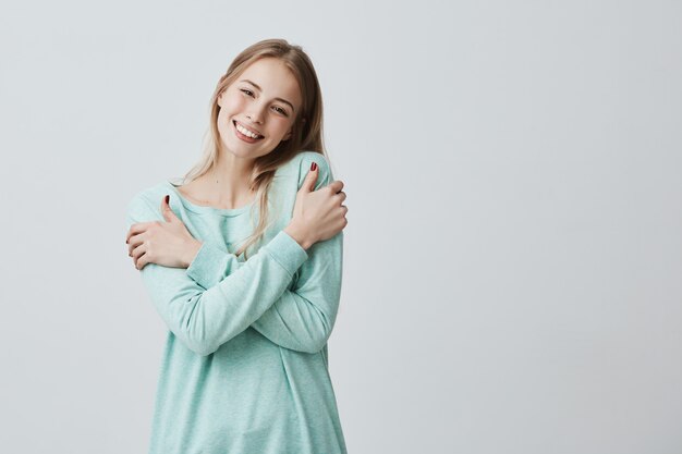 Porträt der reizenden positiven jungen europäischen Frau mit charmantem Lächeln und langen blonden Haaren, die weichen Stoff ihres neuen hellblauen Pullovers genießen, der aufwirft